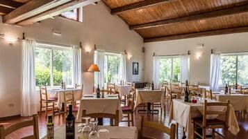 Un restaurant aux poutres apparentes, aux murs blancs et aux grandes fenêtres. Les tables sont dressées avec des bouteilles de vin, des verres et des menus.