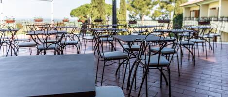 An outdoor patio with tables and chairs, a brick floor, and a view of trees and buildings in the background.