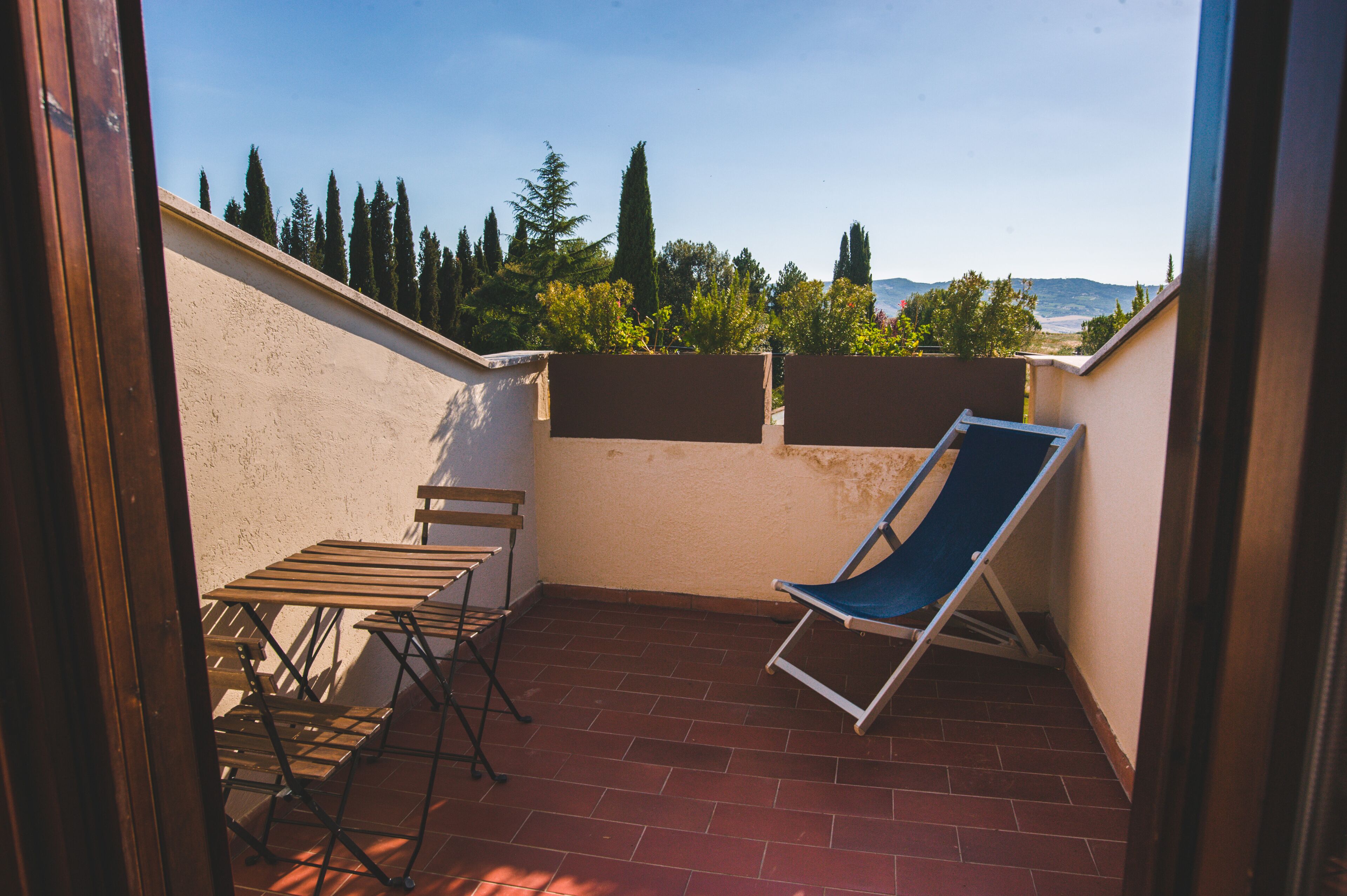 Un pequeño balcón exterior con una mesa y sillas, un sillón azul, y vistas a árboles y colinas.