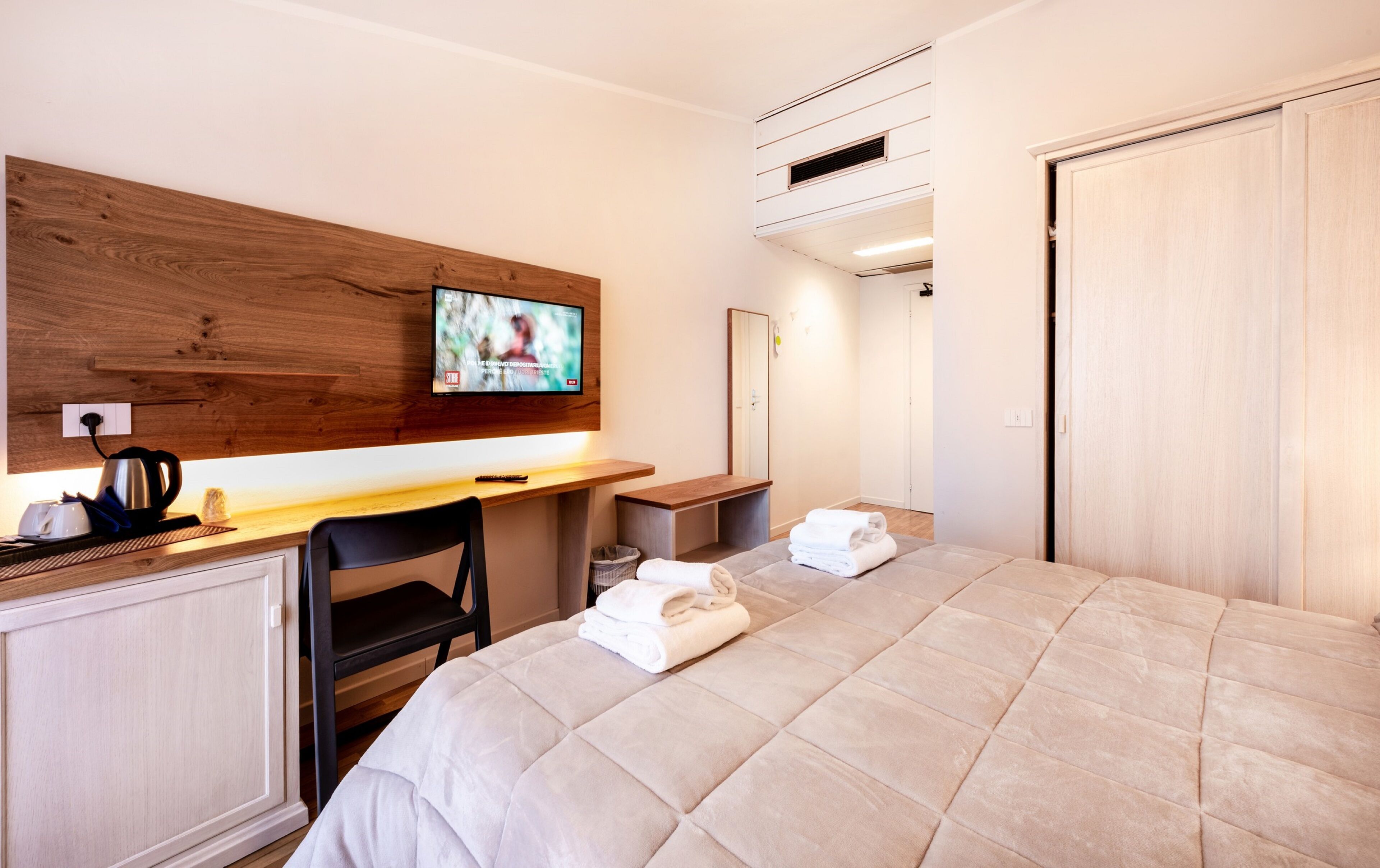 A hotel room with a bed, a desk with a kettle, a chair, a TV, and a wardrobe.