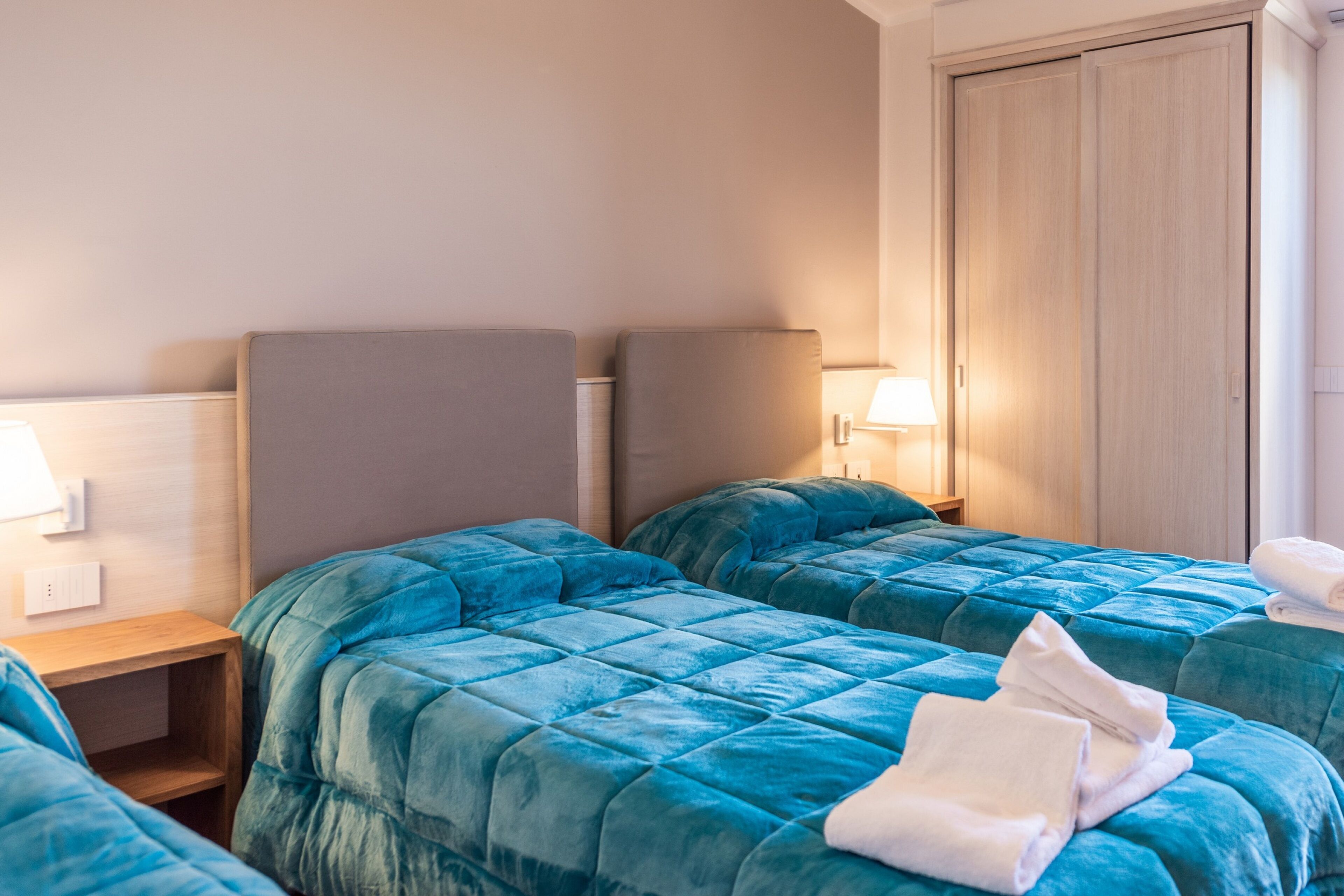 Habitación de hotel con dos camas, cada una con una colcha azul acolchada y un cabecero gris. Se ve una mesita de noche de madera con una lámpara y una toalla blanca doblada sobre la cama.