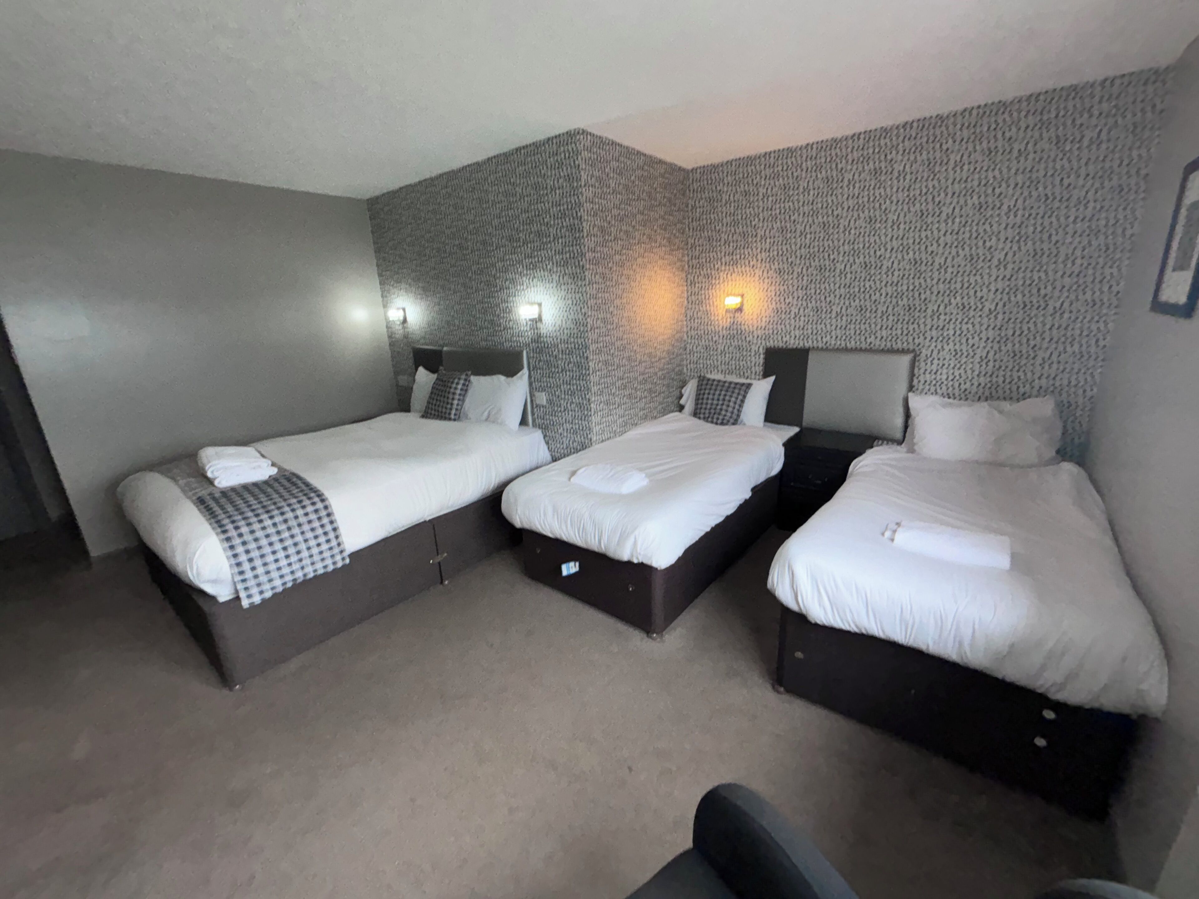 A hotel room with three single beds, each with a pillow and a checkered blanket. The room has a textured wall and two wall-mounted lights.