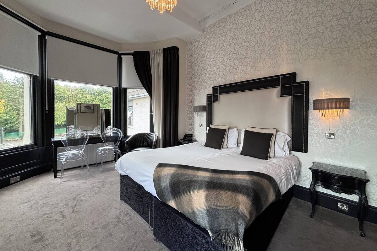 Ein modernes Schlafzimmer mit einem großen Bett, einer Kristallleuchter, Blick durch ein Fenster und einem Schreibtisch mit Lampe.