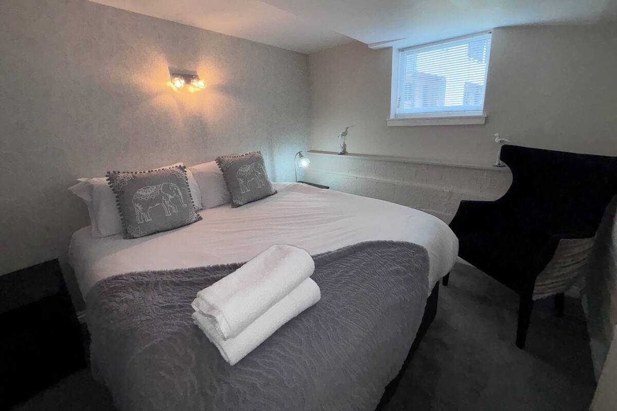 Ein ordentlich bezogenes Bett mit grauer Decke und Kissen, ein kleiner Nachttisch mit Lampe sowie ein Fenster mit Jalousien.