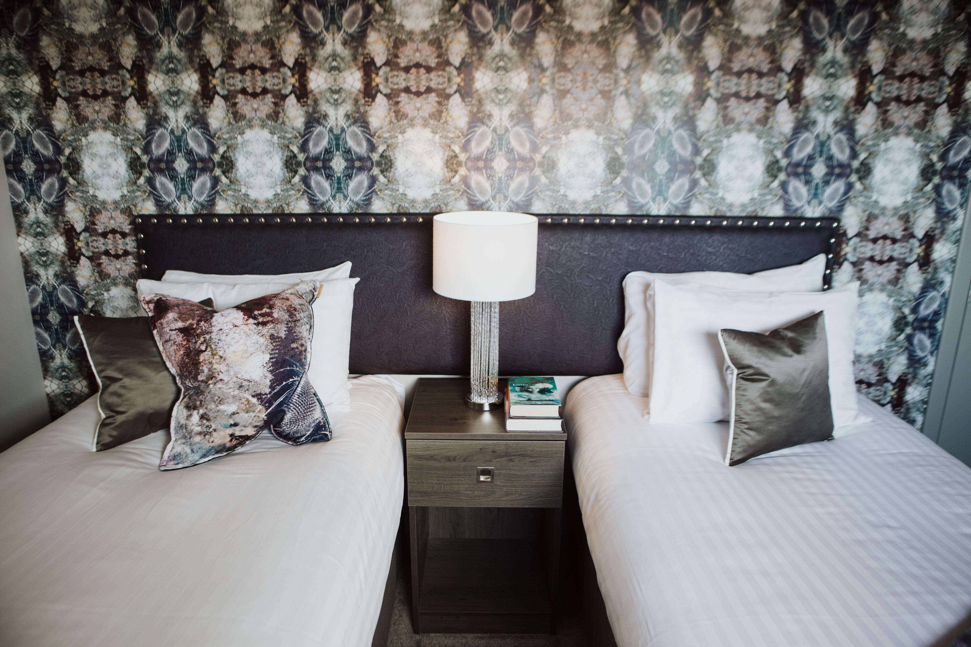A hotel room with two beds, a nightstand with a lamp, and patterned wallpaper.