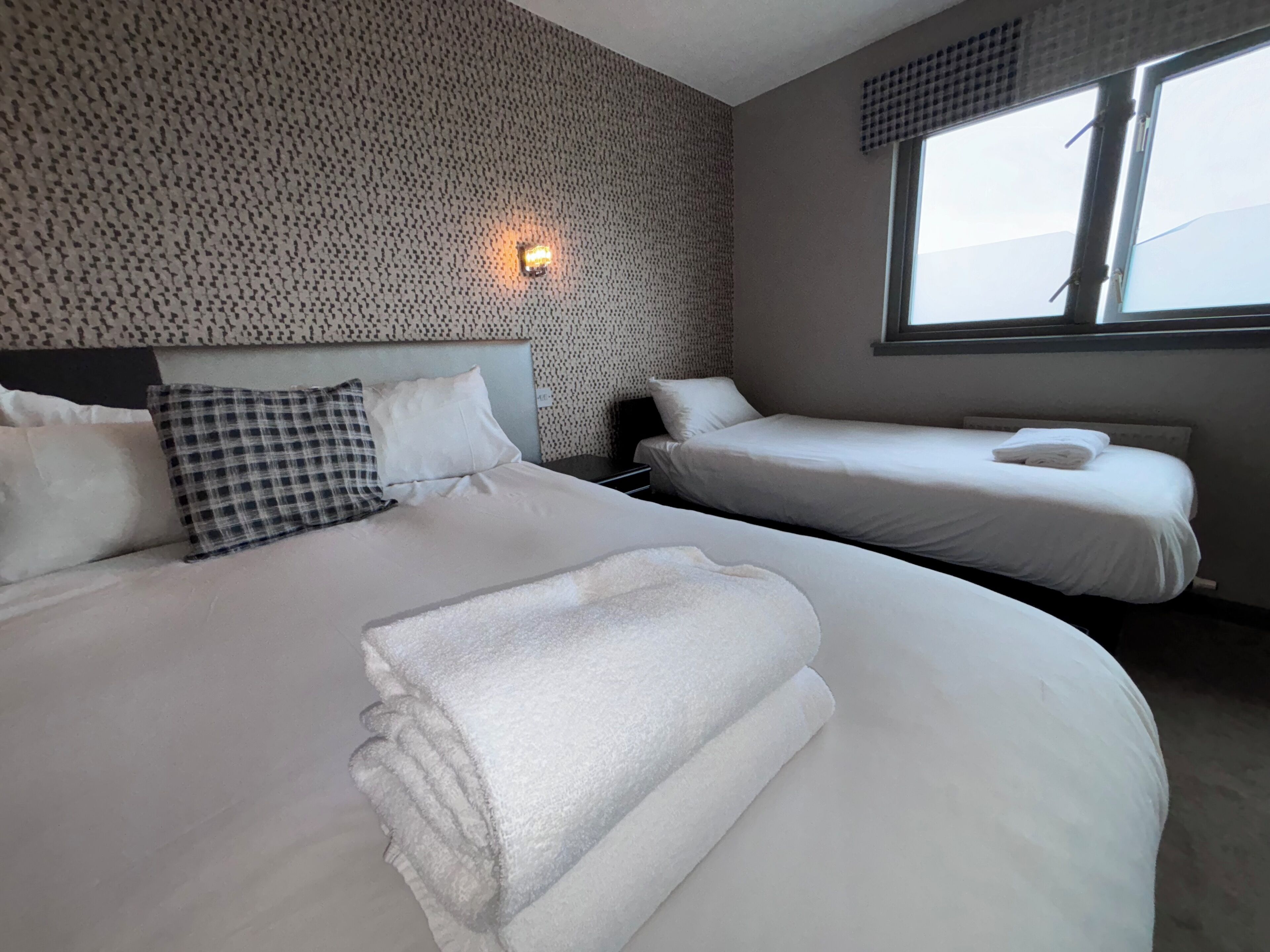 Ein Hotelzimmer mit einem großen Bett, einem kleineren Bett, einem Fenster und einer Wand mit einer strukturierten Oberfläche.