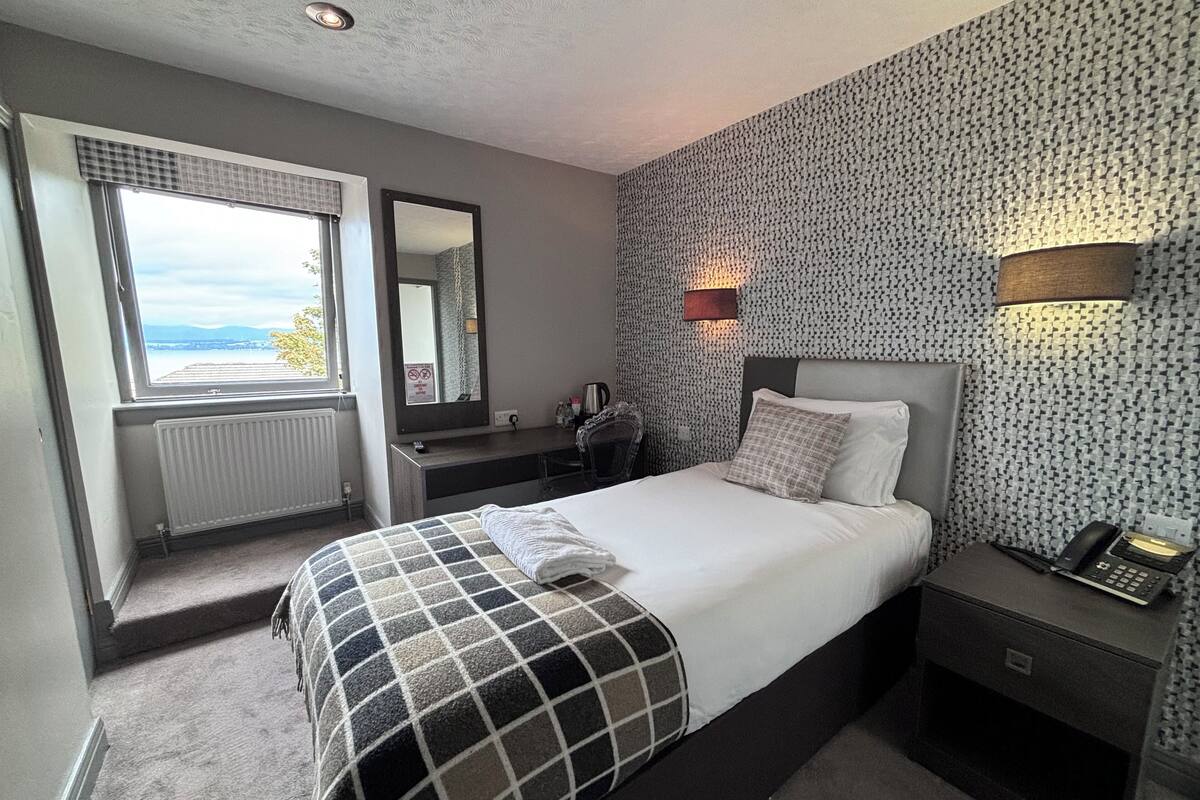 Ein Hotelzimmer mit einem Bett, einem Schreibtisch, einem Stuhl, einem Spiegel, einem Fenster und einer Wand mit ornamentalem Wandpapier.