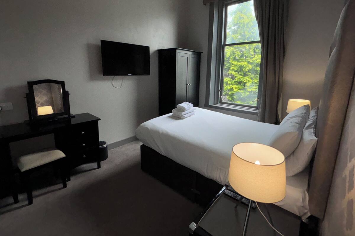 Ein Hotelzimmer mit Bett, Schreibtisch mit Spiegel, Fernseher und einem Fenster mit Vorhängen.
