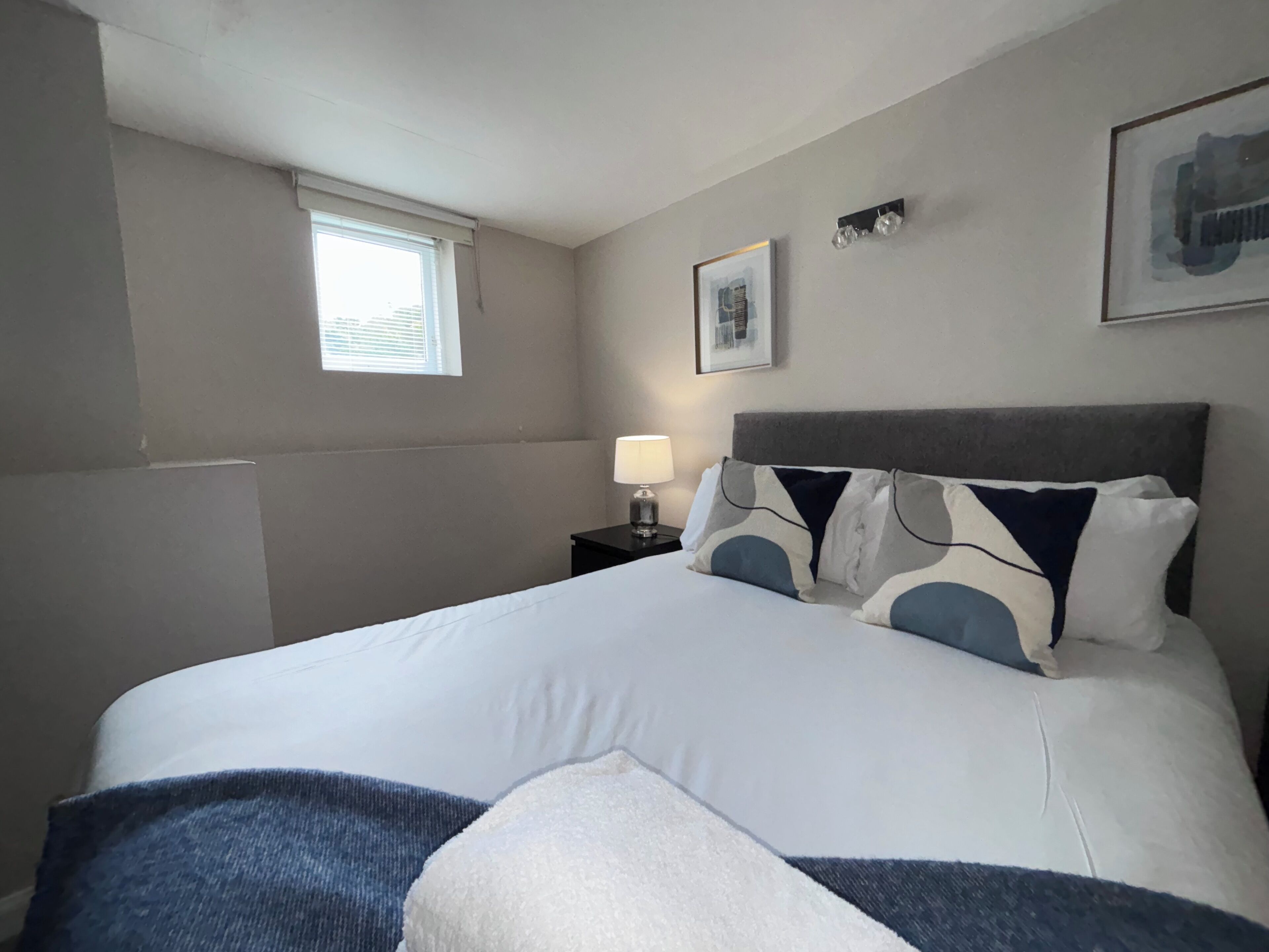 A hotel room with a bed, a nightstand, a lamp, a window, and a framed picture on the wall.