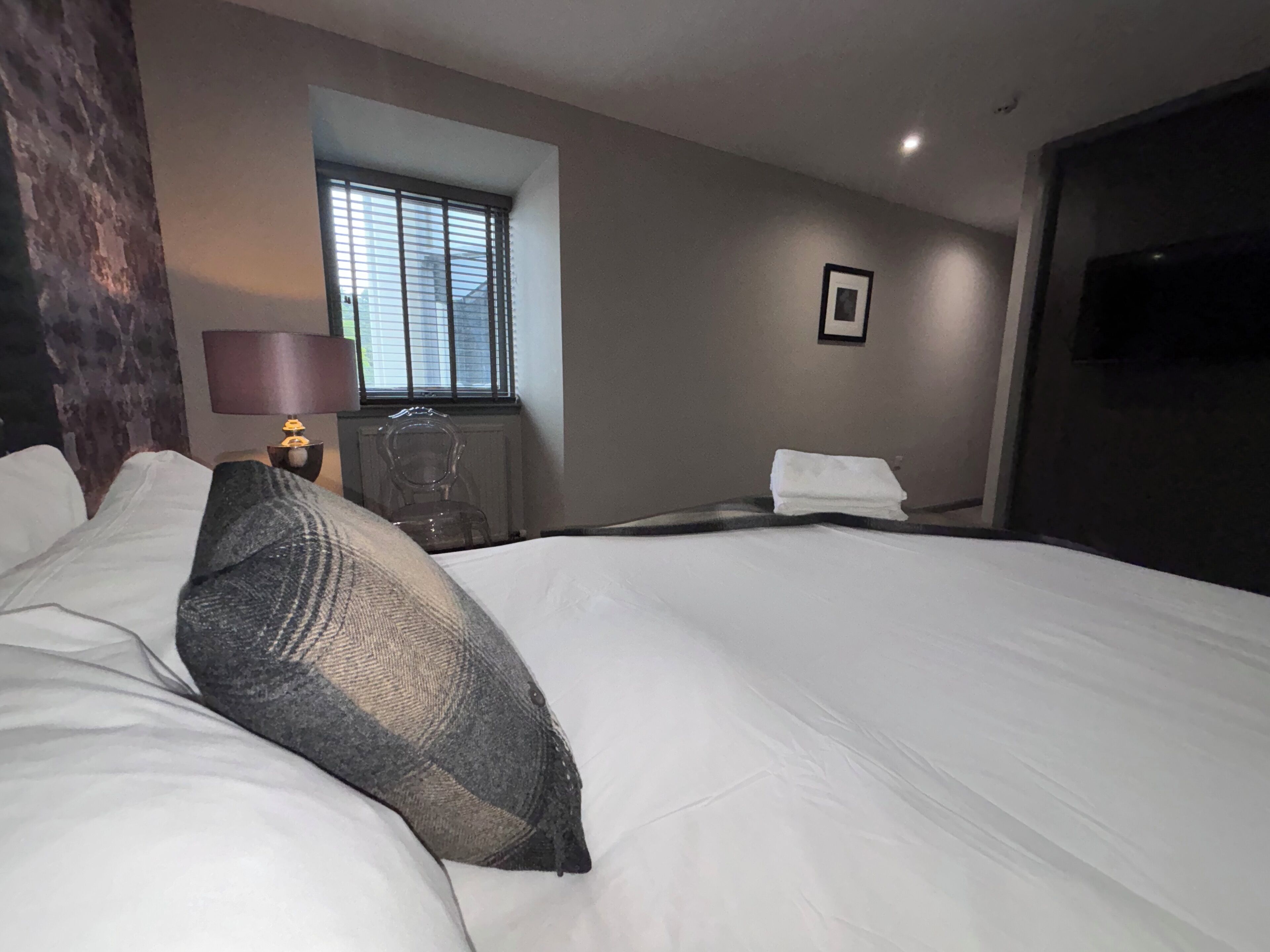 A hotel room with a large bed, a window with blinds, a chair, a radiator, and a framed picture on the wall.