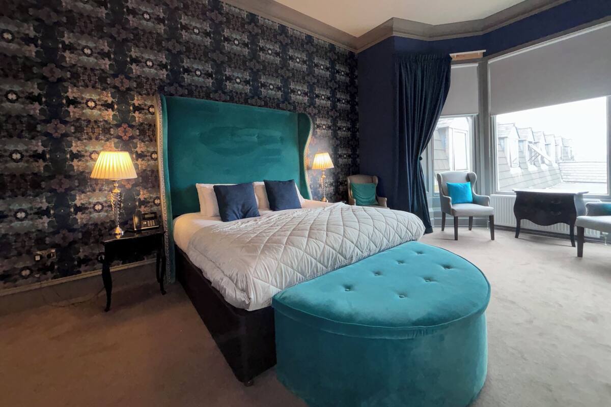 Ein Schlafzimmer mit einem großen Bett, einem salbeengrünem Hocker, einem Sessel, einem Schreibtisch und Blick ins Freie.