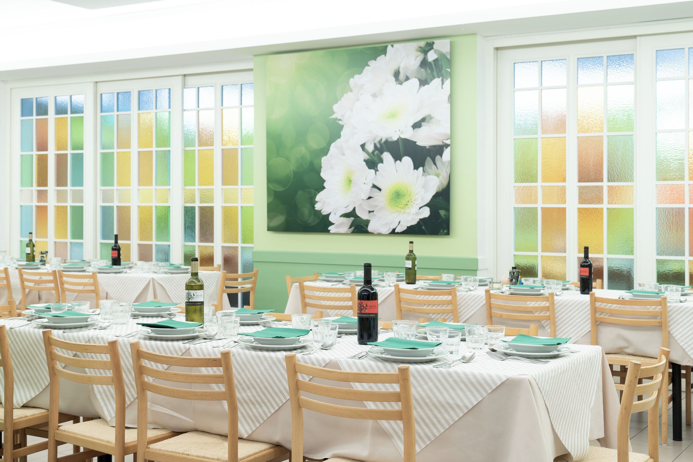 A dining area with tables set for service, featuring a large floral painting on the wall and a view through the windows.