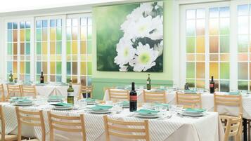 A dining area with tables set for service, featuring a large floral painting on the wall and a view through the windows.