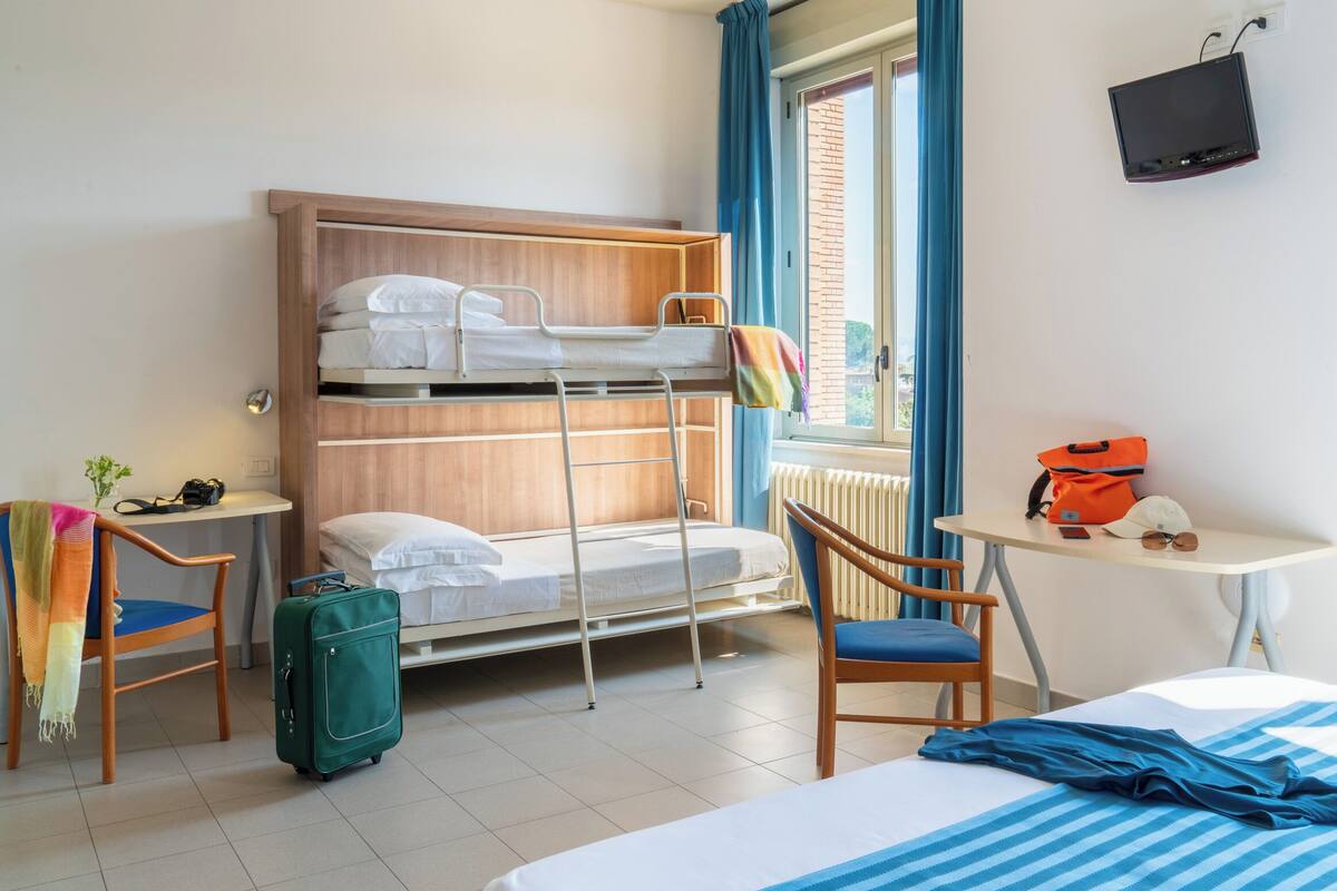 A hotel room with a bunk bed, a desk, a chair, a suitcase, and a television.
