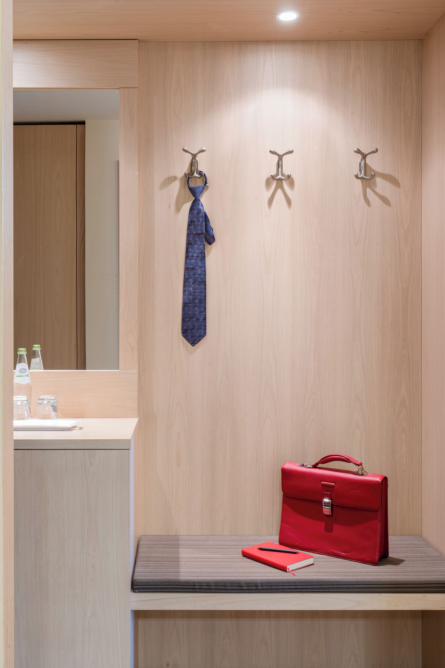 Habitación de hotel con pared de madera, un espejo, un lavamanos y un banco con una maleta roja y un cuaderno.