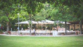 Un patio al aire libre con mesas y sombrillas, rodeado de árboles y plantas en macetas.