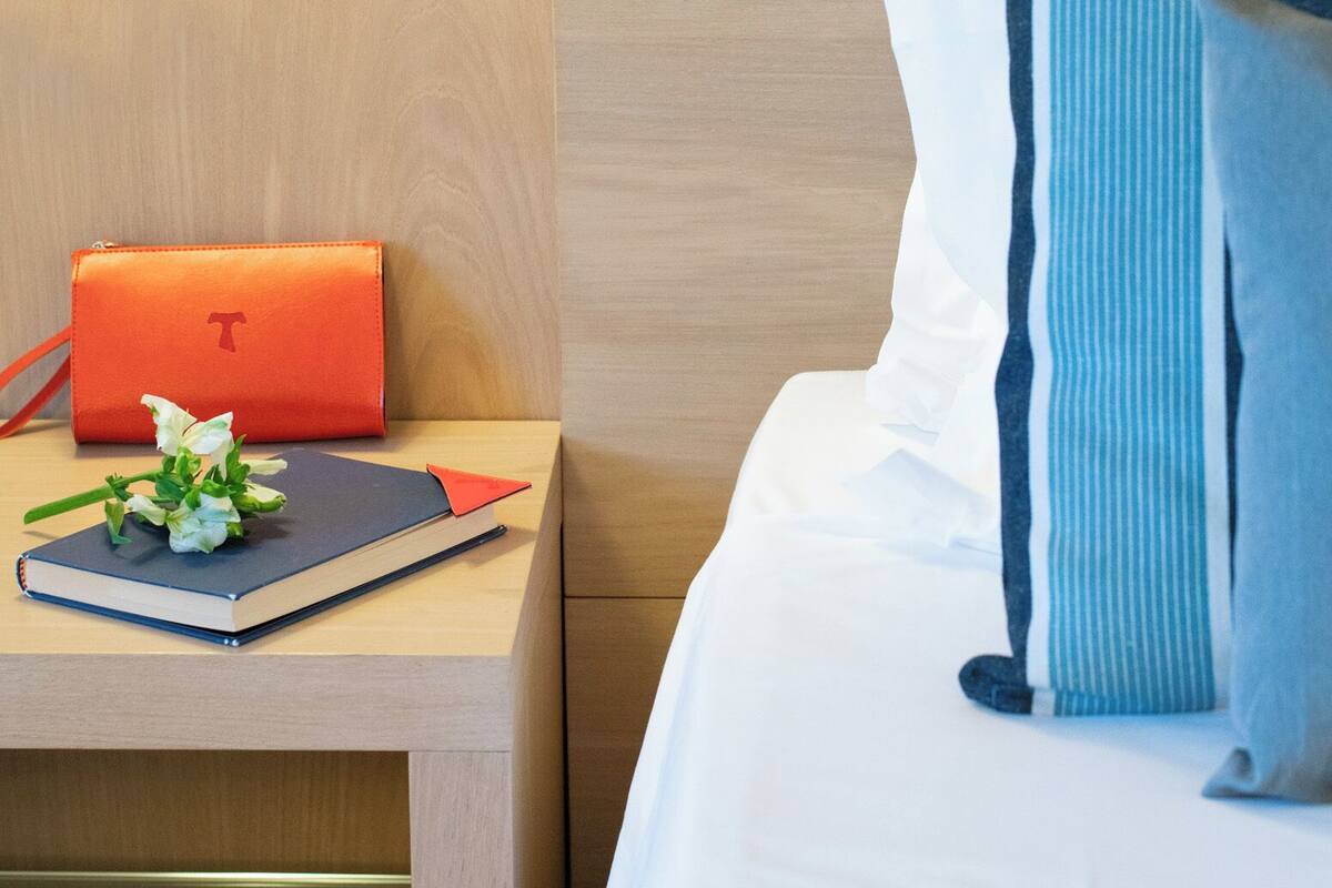 A neatly made bed with a striped pillow and a bedside table with a book, a flower, and a red bag.