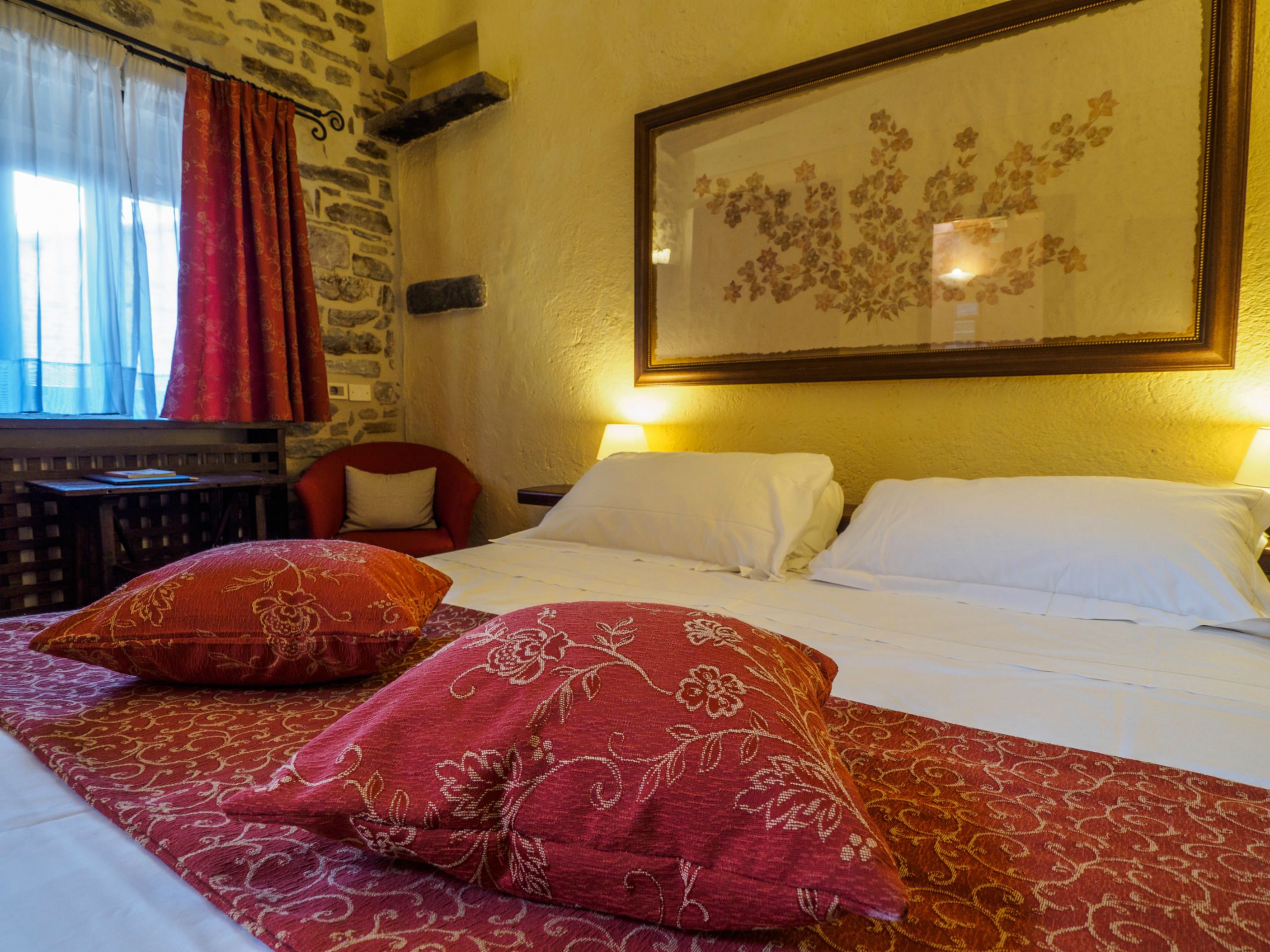Habitación de hotel con cama, dos almohadas, una silla, una mesa y un cuadro floral en la pared.