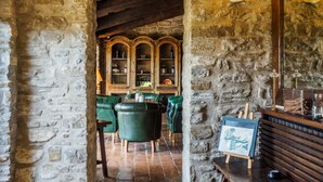 Un interior rústico con paredes de piedra, vigas de madera y sillones de cuero verde. Se observan armarios de madera con botellas y un cuadro en un escritorio.