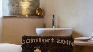 Un asciugamano con la scritta "comfort zone", una lozione e una candela accesa su un tavolo in una stanza con un lavandino e un elemento decorativo a parete.