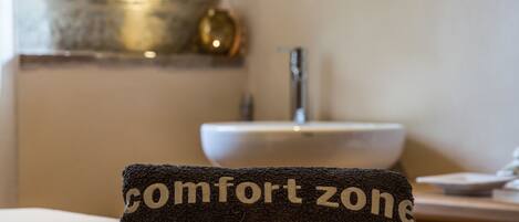Een handdoek met de tekst 'comfort zone', een fles lotion en een brandende kaars op een tafel in een kamer met een wastafel en een decoratief wandobject.