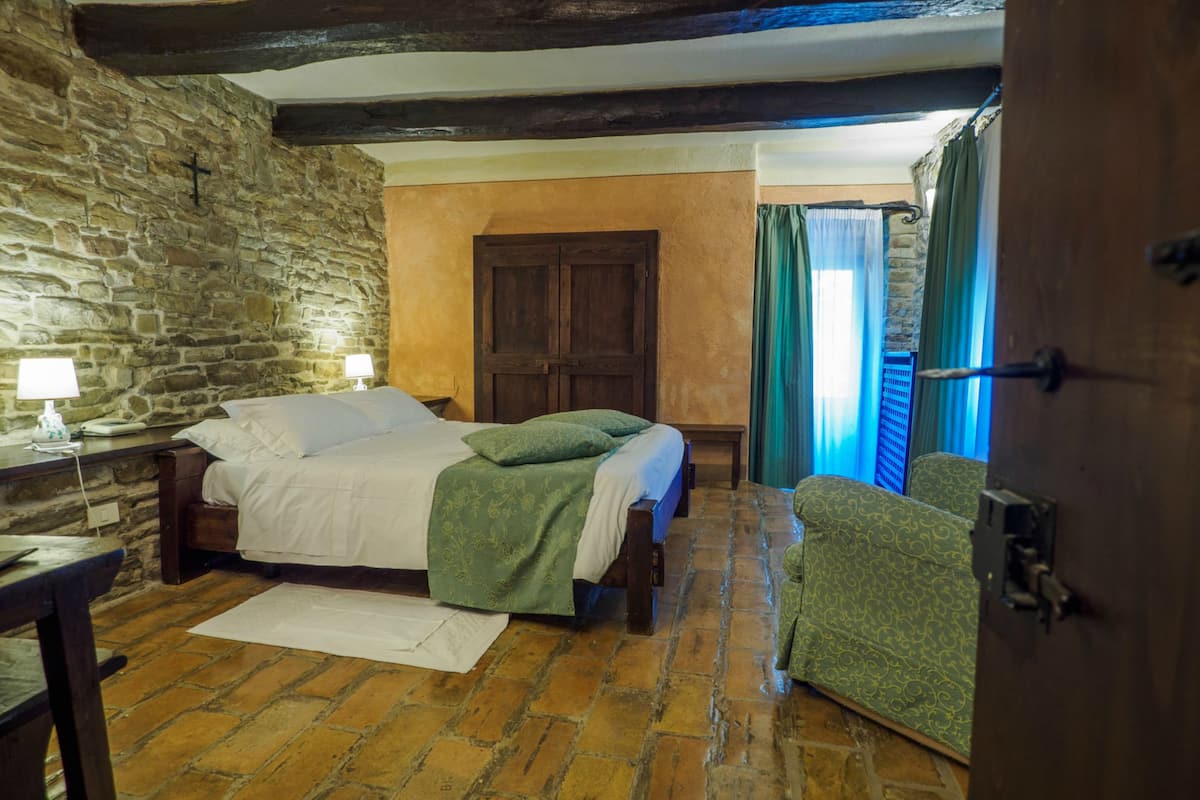 Ein traditionell eingerichteter Raum mit Steinwand, Holztür, einem Bett mit grüner Bettwäsche, einem Sessel und einem kleinen Tisch mit Lampe.