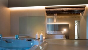 Un baño moderno con una bañera grande, dos velas y un espejo que refleja un dormitorio con una cama y una mesita de noche.