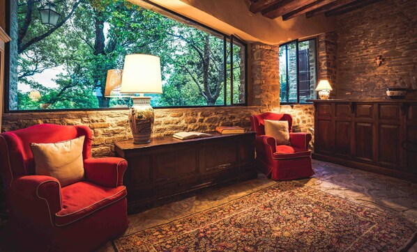 Un coin lecture confortable avec deux fauteuils rouges, une grande fenêtre donnant sur les arbres, un mur en pierre et un tapis à motifs.