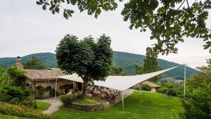 Una casa de piedra con un patio, mobiliario exterior y un gran árbol que da sombra.