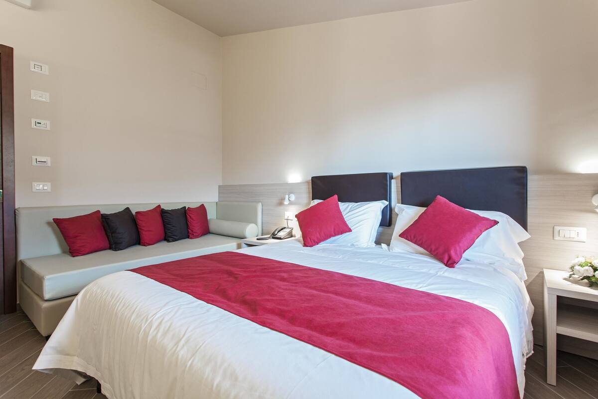 Una habitación de hotel con una cama grande, una mesita de noche con lámpara, un mueble auxiliar y un sofá con cojines rojo y negro.