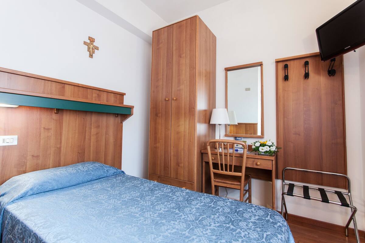 Un dormitorio con cama, armario de madera, escritorio, silla y televisor montado en la pared.