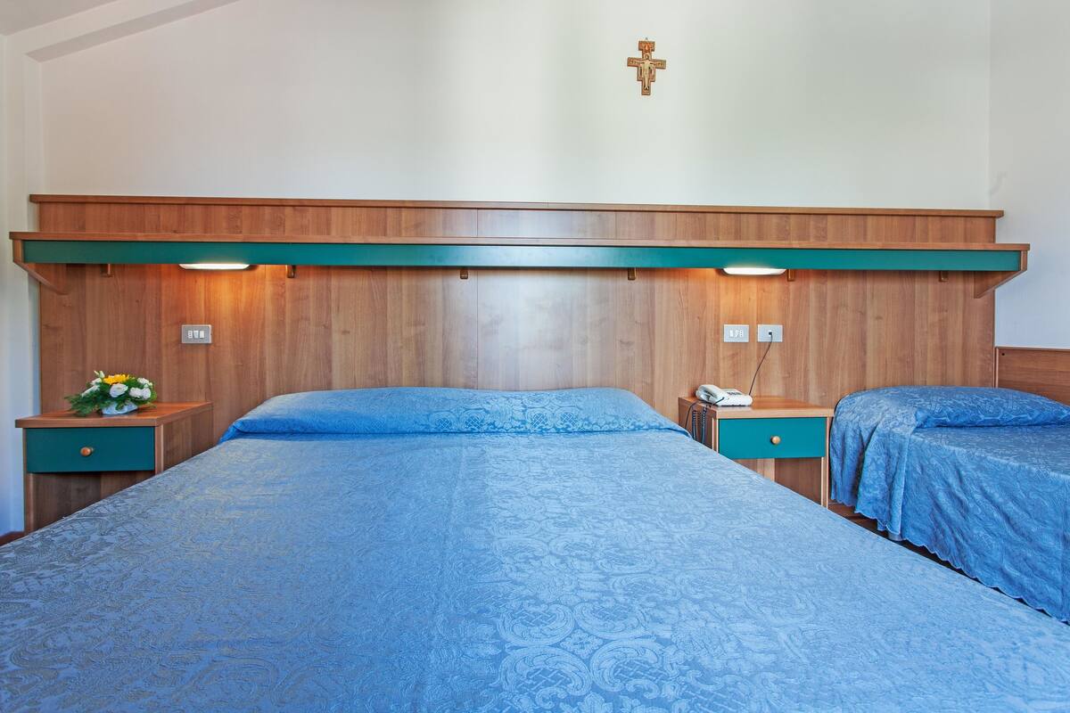 Una habitación de hotel con una cama grande, dos mesitas de noche, un cabecero de madera y una cruz colgada en la pared.