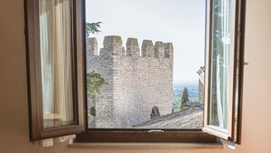 A room with a window overlooking a stone wall, a table with a bottle and fruit, and a curtain partially drawn. - Hotel San Pietro (Assisi)