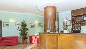 A reception area with a wooden reception desk, a red sofa, a potted plant, and framed pictures on the walls. - Hotel San Pietro (Assisi)
