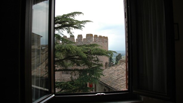 A view through a window of a stone building with a large tree in the foreground and a castle-like structure in the background. - Hotel San Pietro (Assisi)