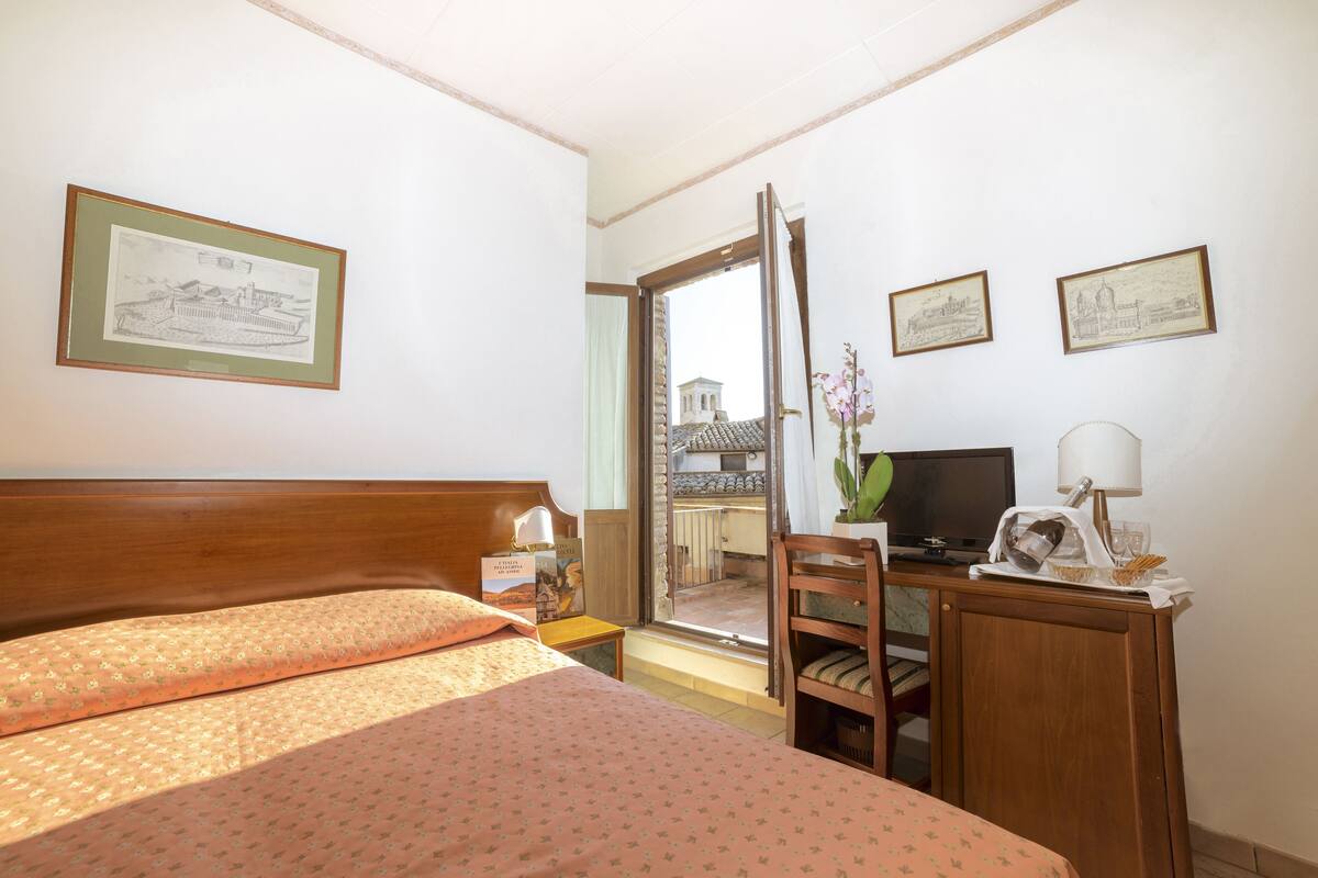 A hotel room with a bed, a desk with a television, a chair, and a window with a view of a building.