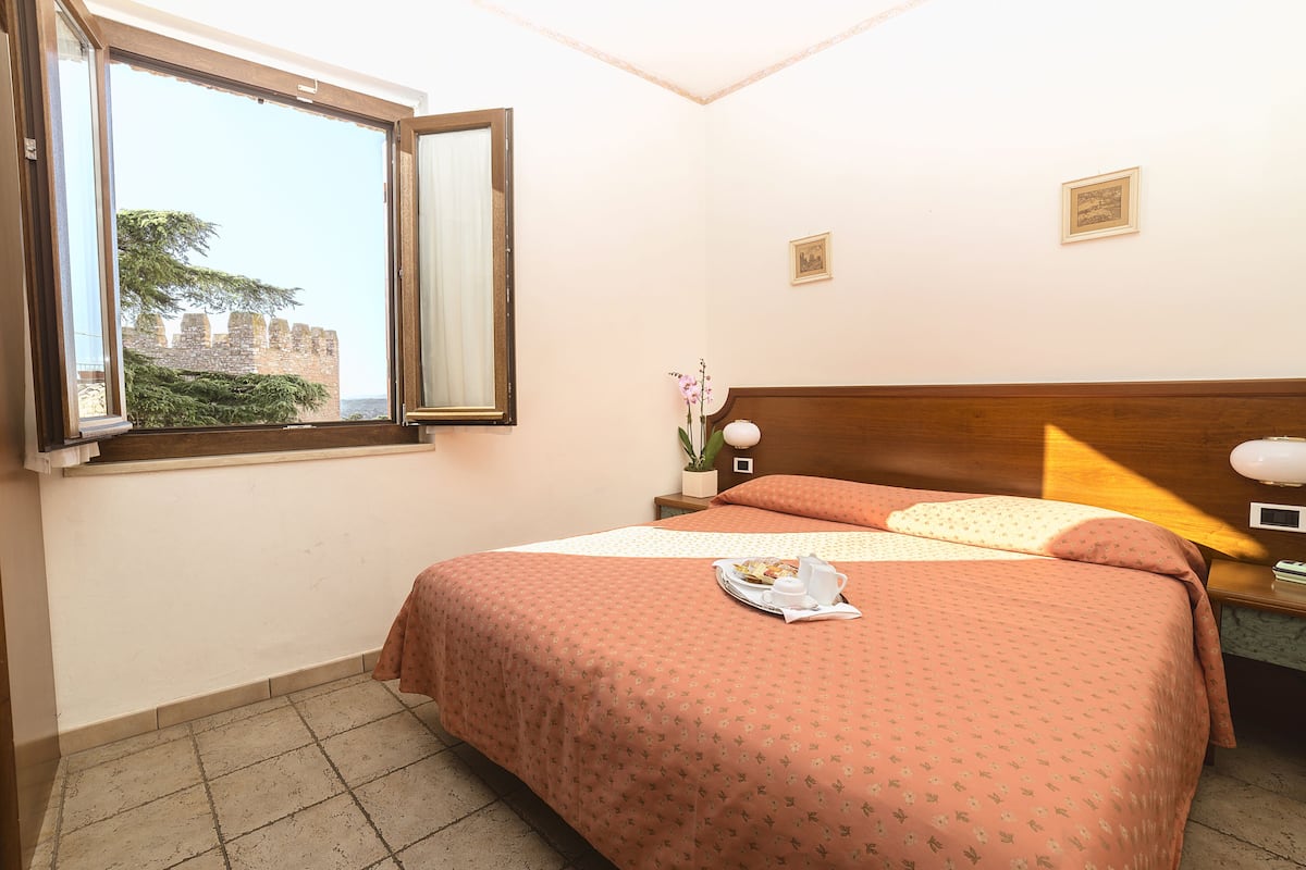 A hotel room with a large bed, a nightstand, a window with an open view, and a small potted plant.