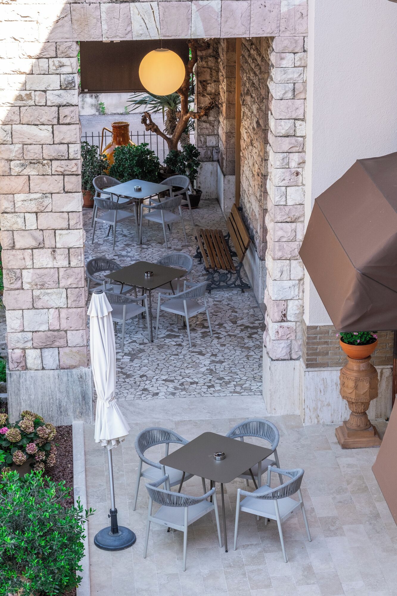 Un patio al aire libre con muros de piedra, varias mesas y sillas, una lámpara colgante y plantas en macetas.