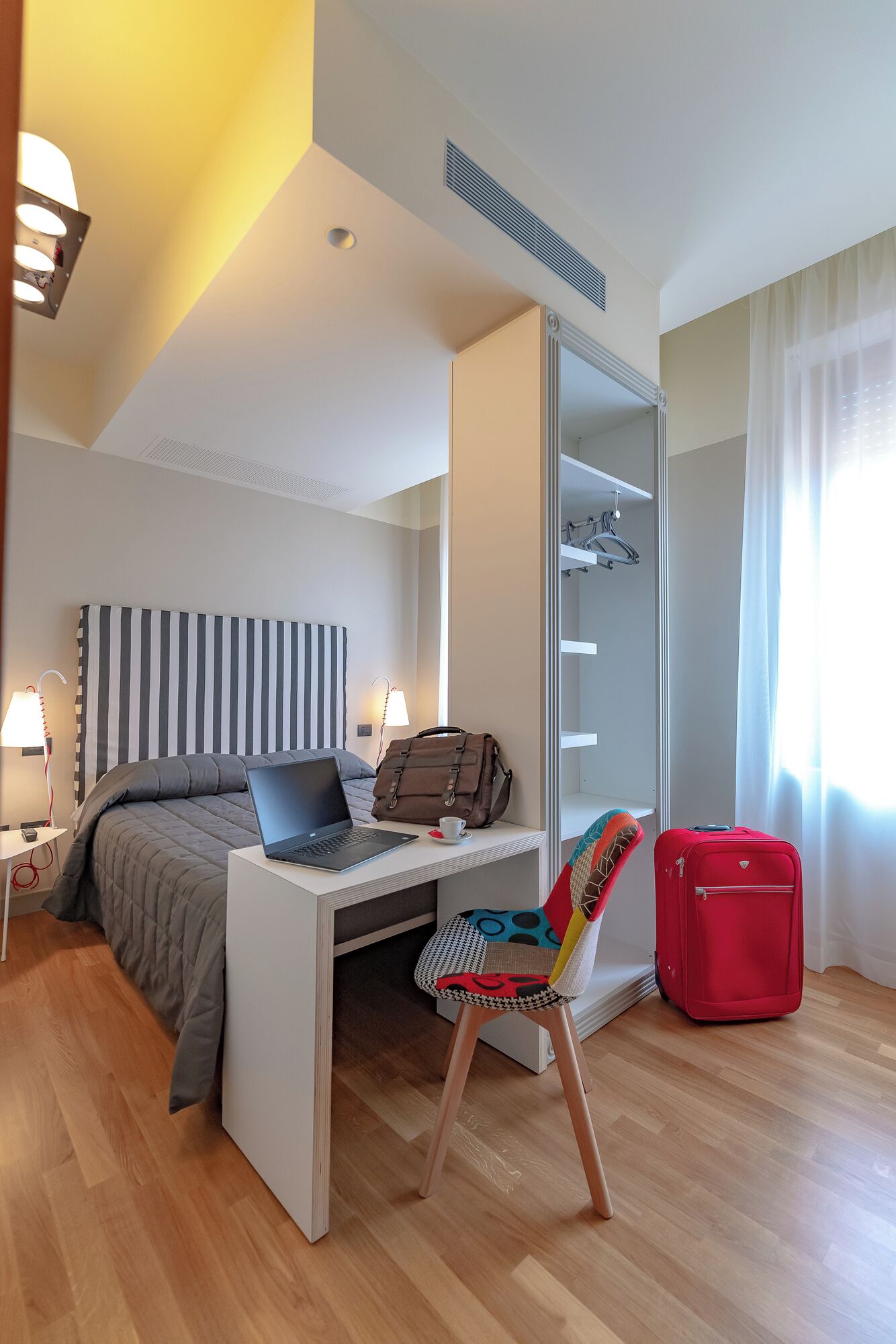 A modern hotel room with a bed, a desk, a chair, a red suitcase, and a wardrobe.