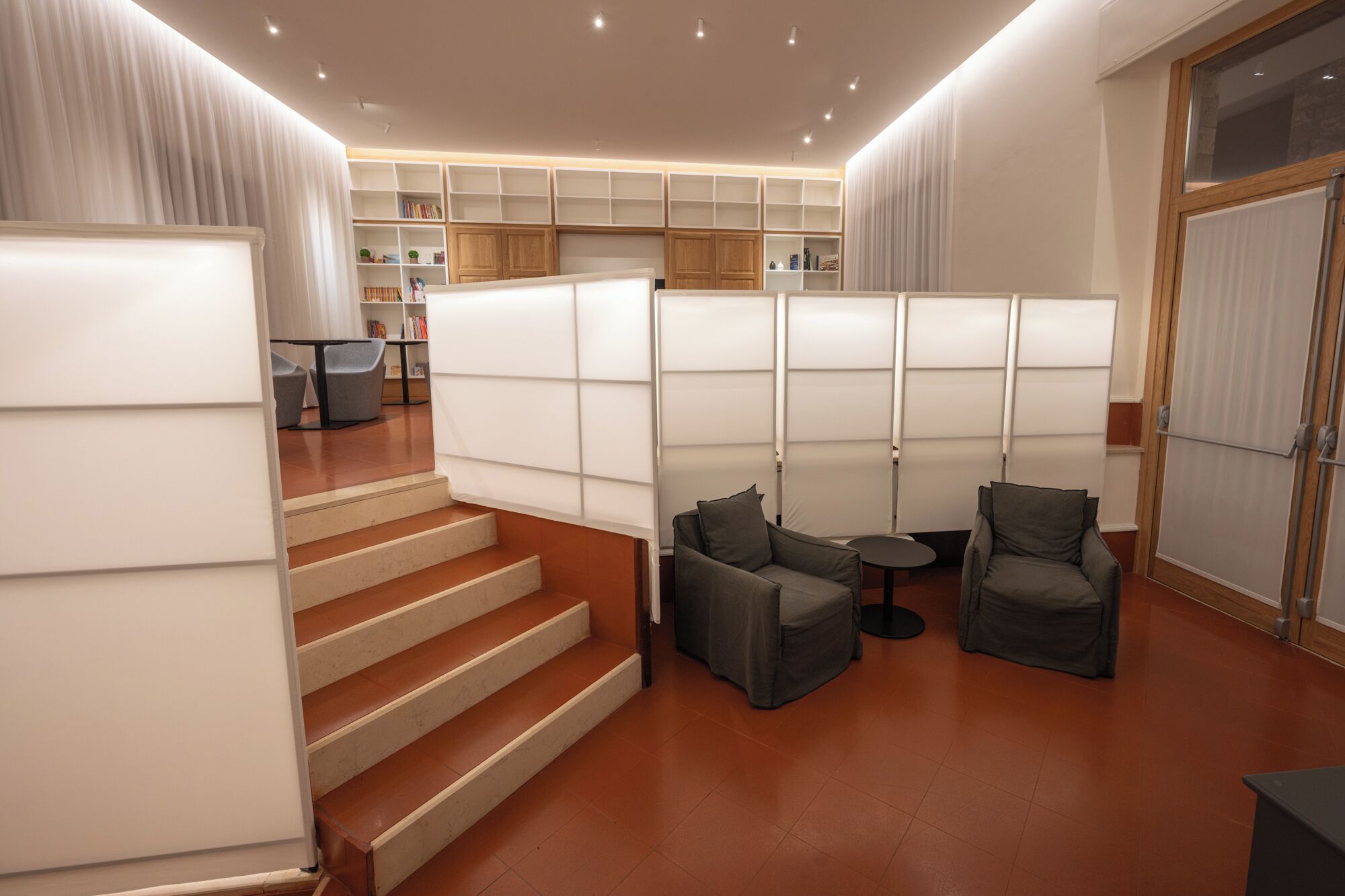 A modern library with tiered bookshelves, comfortable seating, and a staircase.