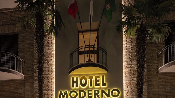 The entrance of Hotel Moderno with a modern design, featuring a brick facade, palm trees, and a prominent illuminated sign. - Hotel Moderno (Assisi)