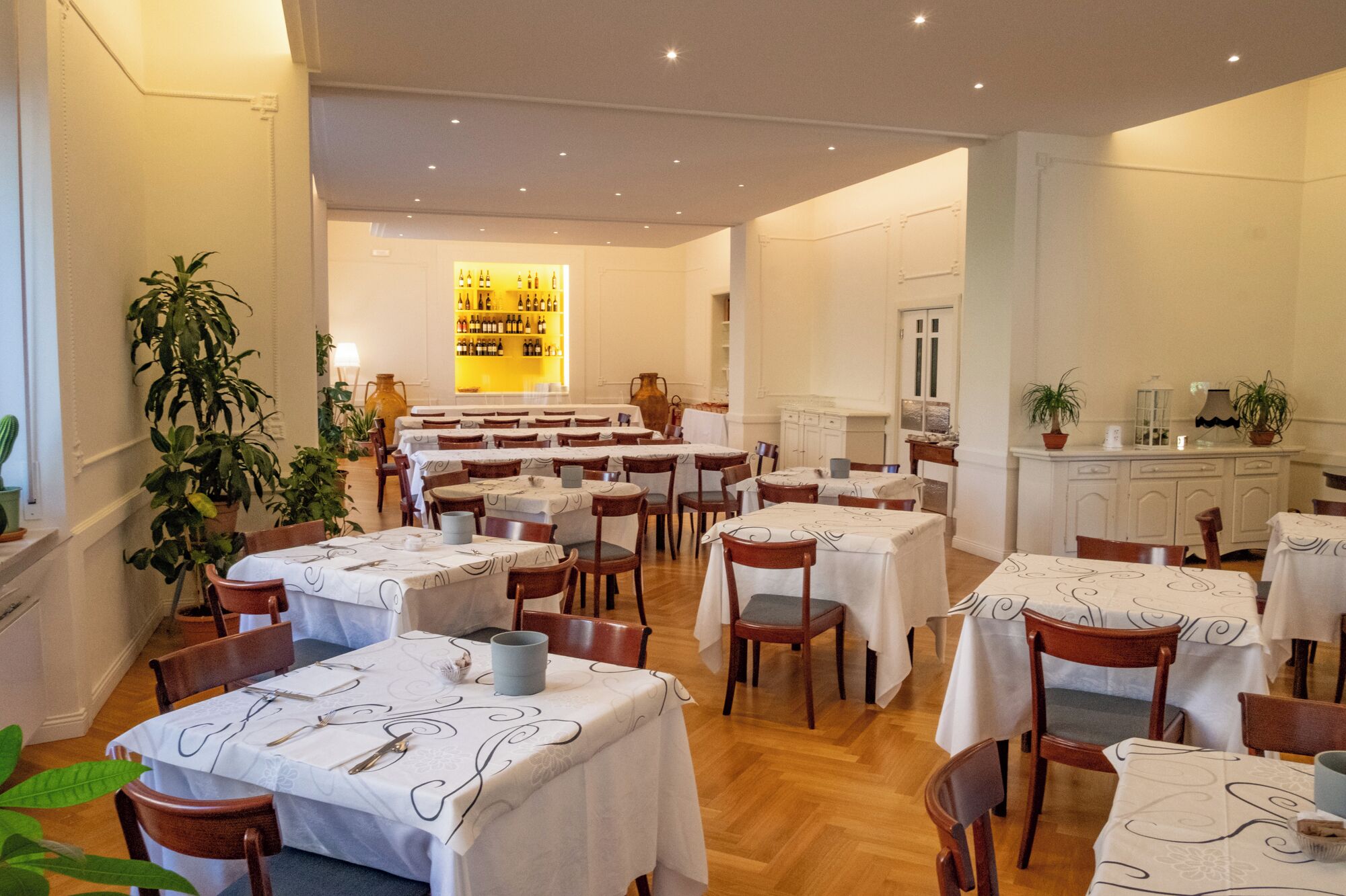 Un restaurante con varias mesas y sillas, una exhibición de vinos y plantas en macetas.