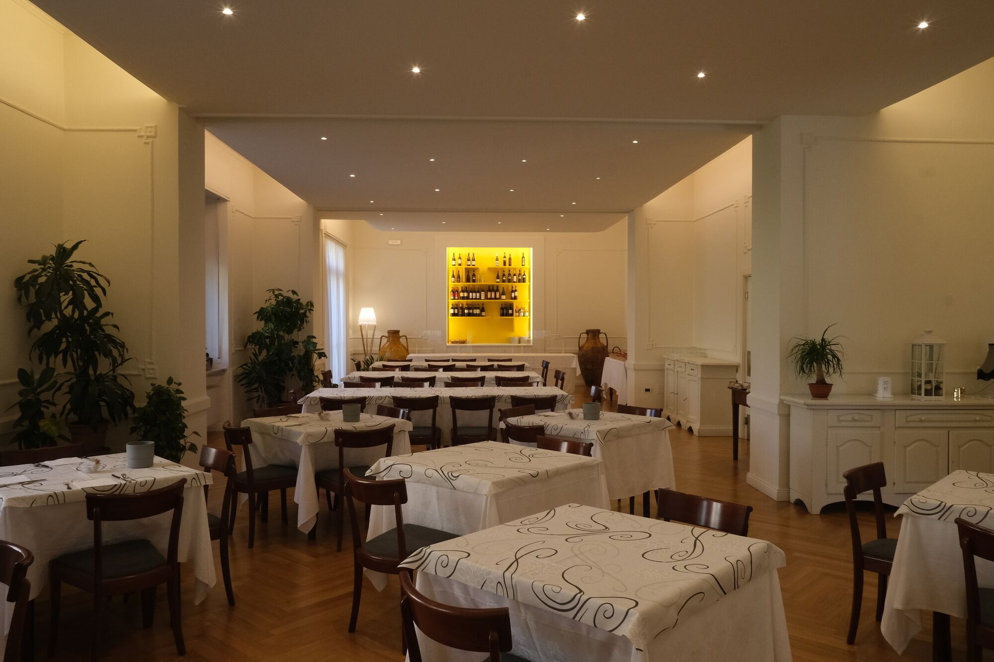 A dining area with multiple tables and chairs, a bar with shelves of bottles, and potted plants.