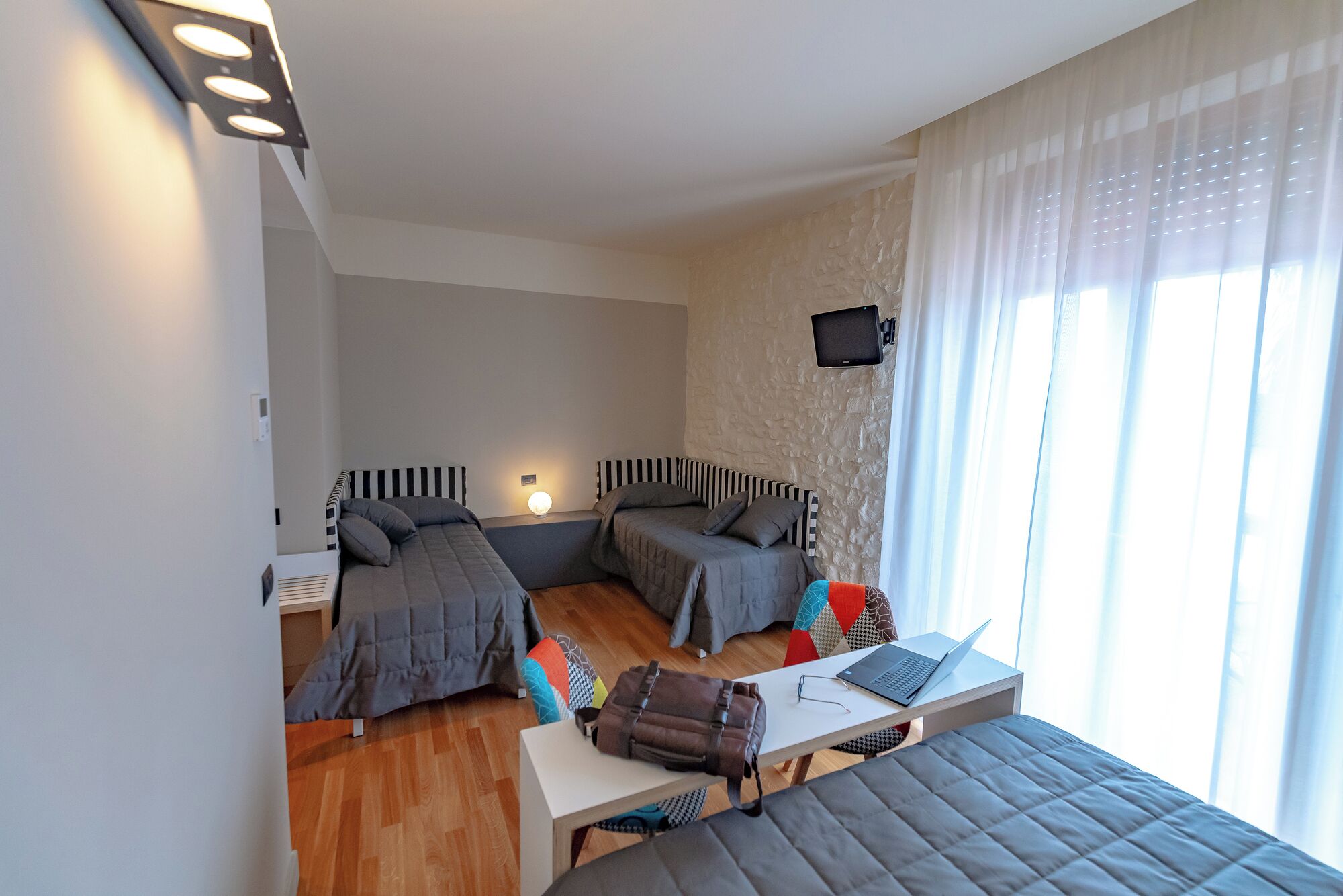 Un dormitorio moderno con dos camas, un escritorio con una computadora portátil y un televisor en la pared.