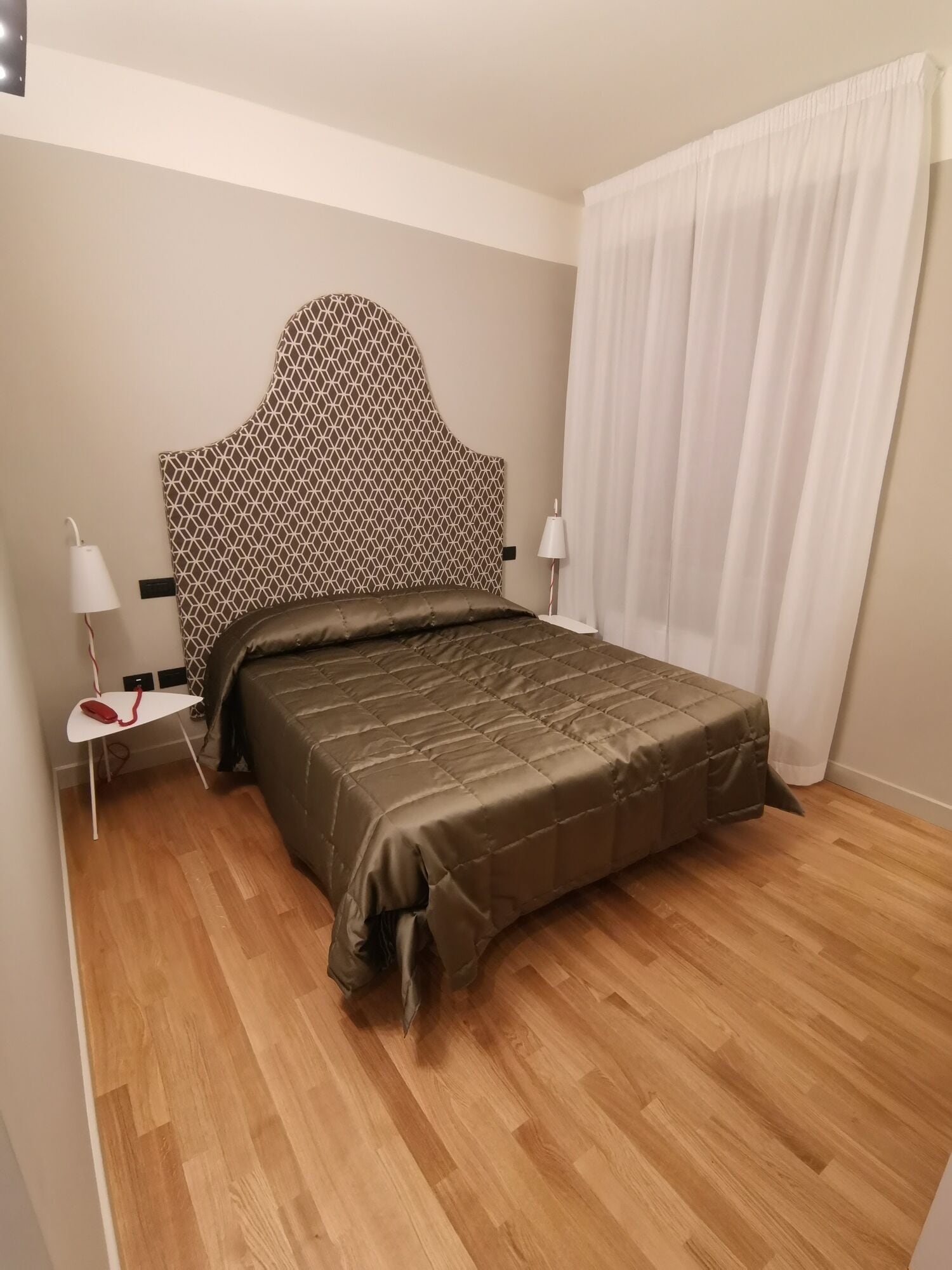 A bedroom with a patterned headboard, a bed with a brown quilt, a bedside table with a lamp, and a white curtain.