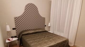 A bedroom with a patterned headboard, a bed with a brown quilt, a bedside table with a lamp, and a white curtain.