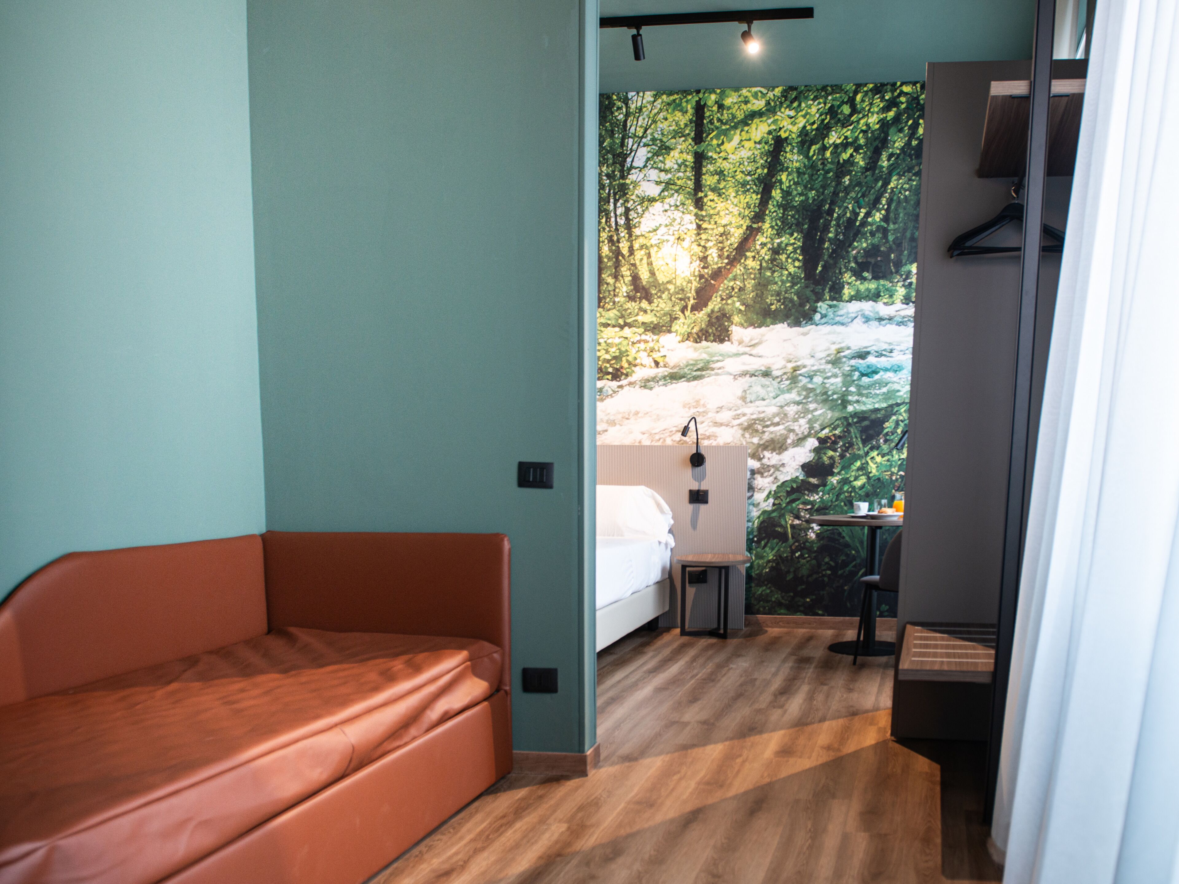 A room with a bed, a red sofa, a small table, and a view of a forest through a window.