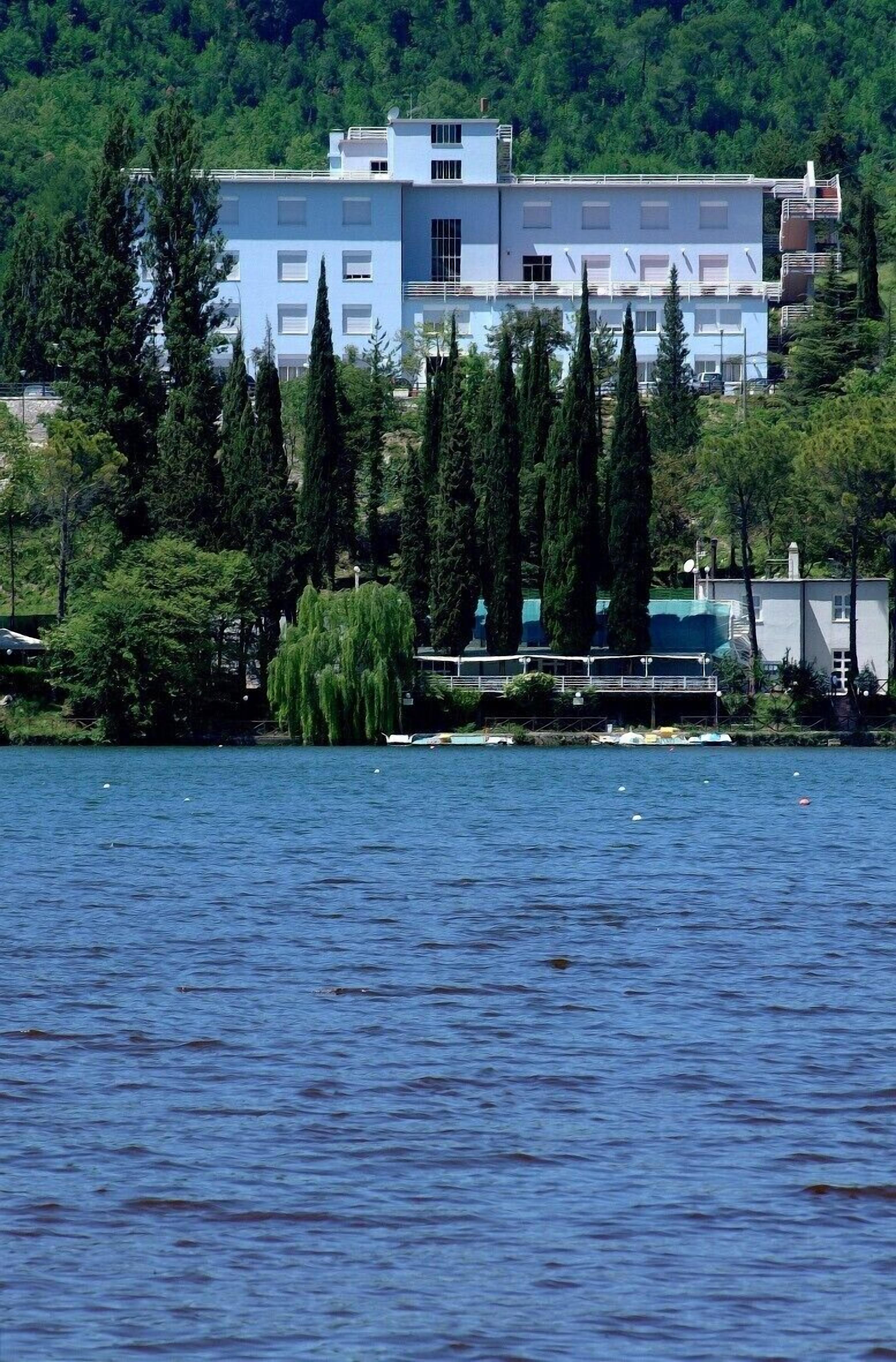 A modern building with a light blue facade, surrounded by tall trees and a body of water.