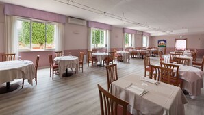 A spacious dining area with multiple tables and chairs, large windows with curtains, and a clean, light-colored floor.