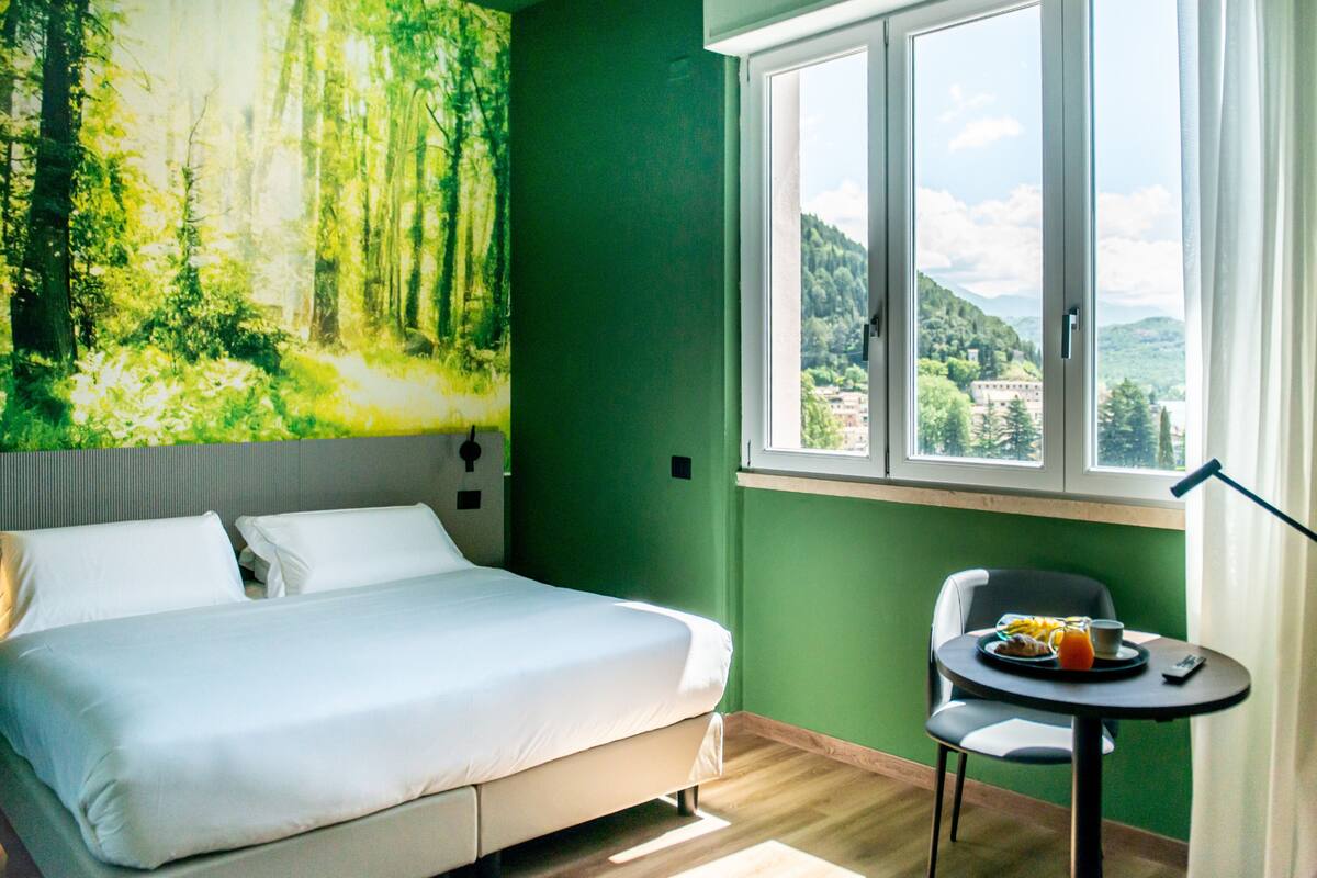 Ein Hotelzimmer mit Bett, kleinem Tisch mit Frühstück, Stuhl, Fenster mit Blick auf Wald und Berge sowie einer grünen Wand.